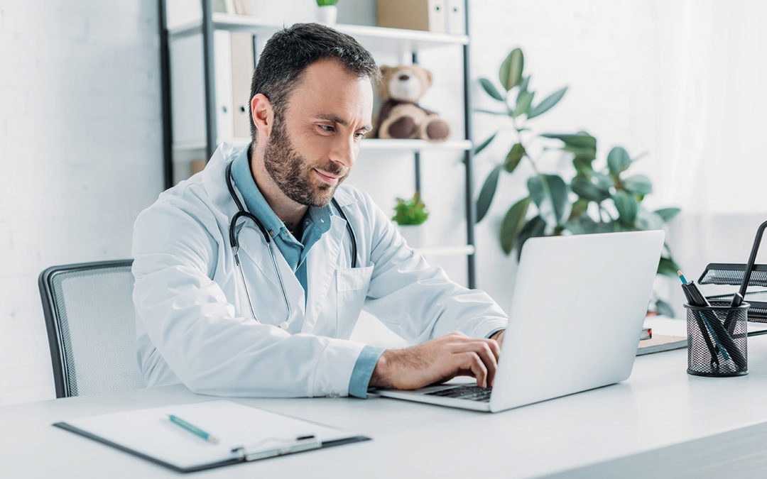 positive doctor sitting at workplace and using laptop - Implicity